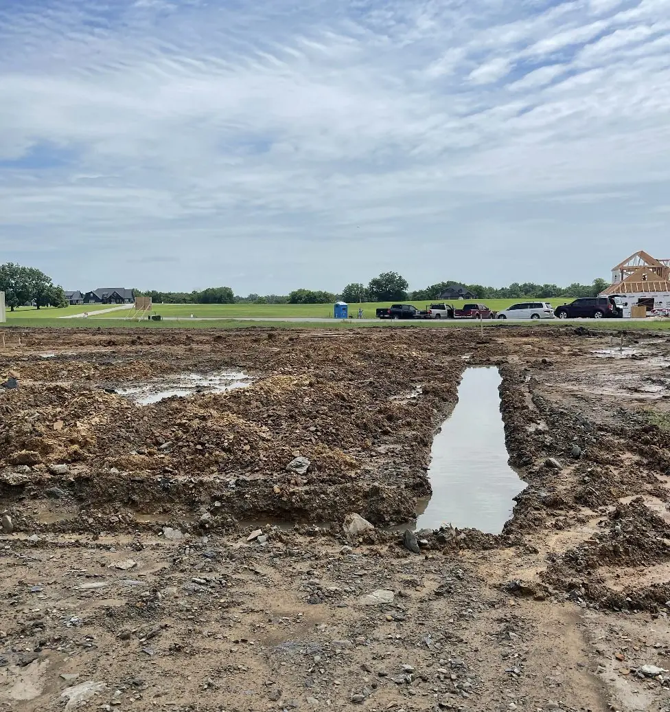 Freshly graded lot with a low spot holding rainwater, new houses beyond