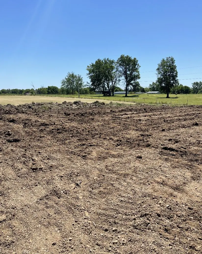 Bare dirt graded across a field with a stand of trees behind