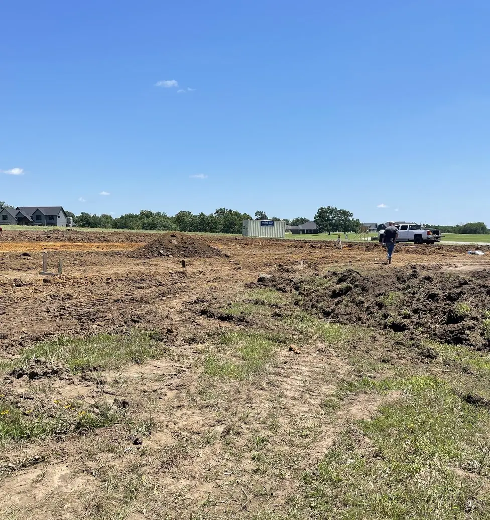 Site graded to dirt with stakes set out and a pickup beyond